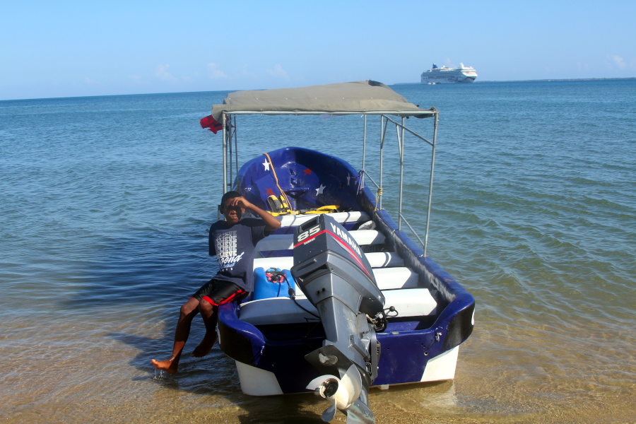 A calm ocean view with a young man relaxing by a boat, symbolizing the grounding and release that somatic healing for anxiety brings to both body and mind. Young man resting beside a small boat on the shore with calm water and a cruise ship in the distance, reflecting the peace that somatic healing for anxiety can restore.